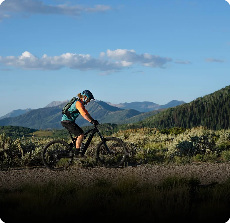 Person in mountain bike gear riding an electric mountain bike on a dirt trail with mountains in the background.